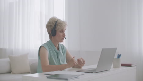 female-online-consultant-is-talking-with-client-by-headphones-with-microphone-looking-at-screen-of-laptop-working-from-home-distant-work